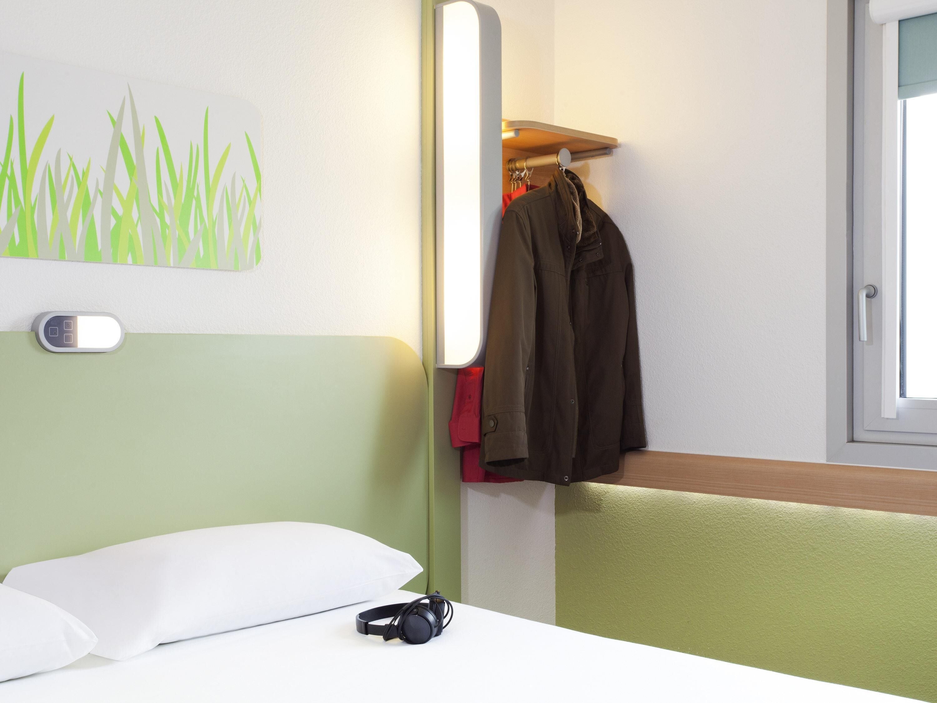 A hotel room with a bed, a wall-mounted light, a coat rack with clothes, and a window.