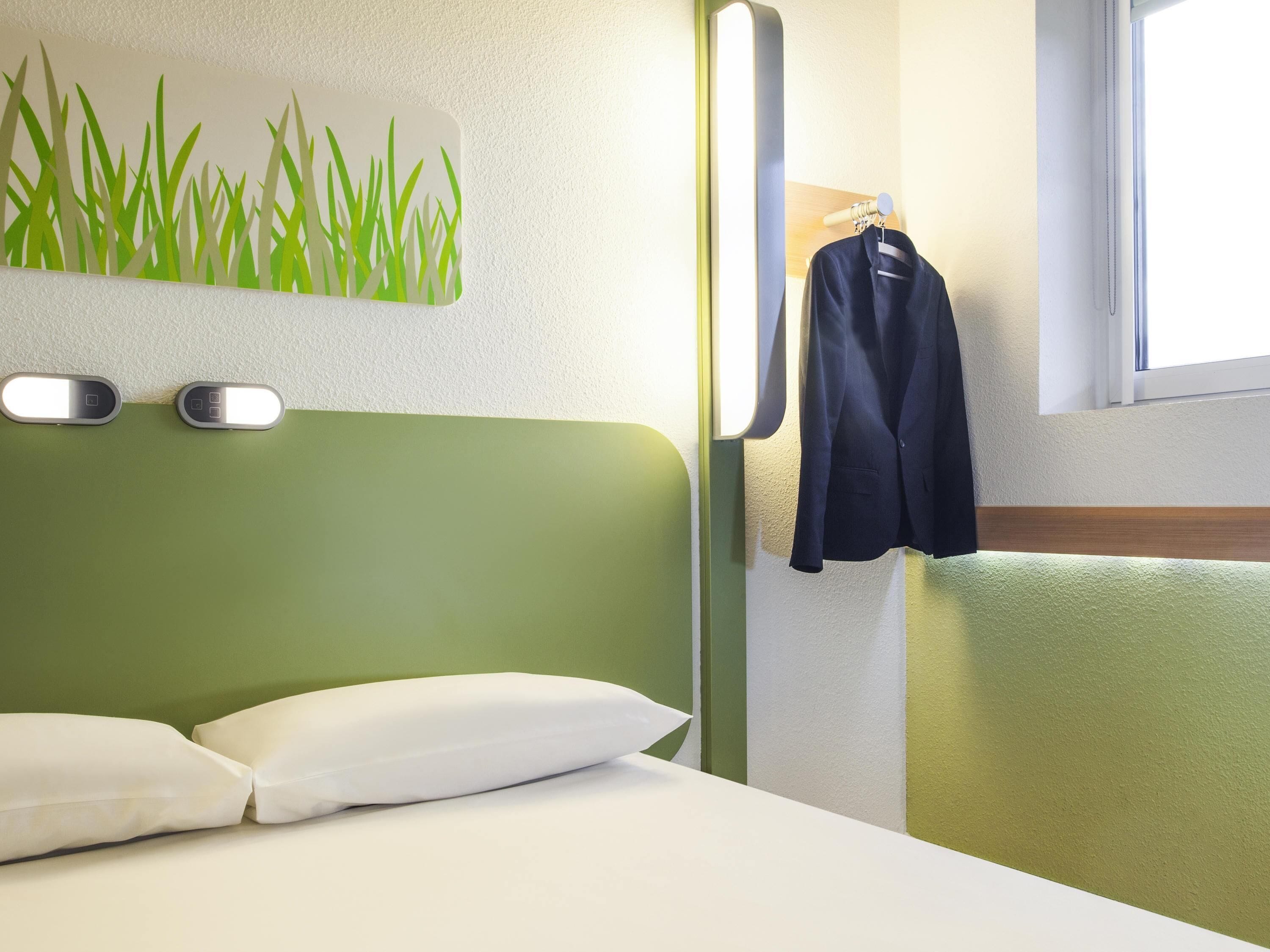 Una habitación de hotel con cabecera verde, almohadas blancas, una lámpara fijada a la pared, un abrigo colgado de un perchero y una ventana con luz natural.