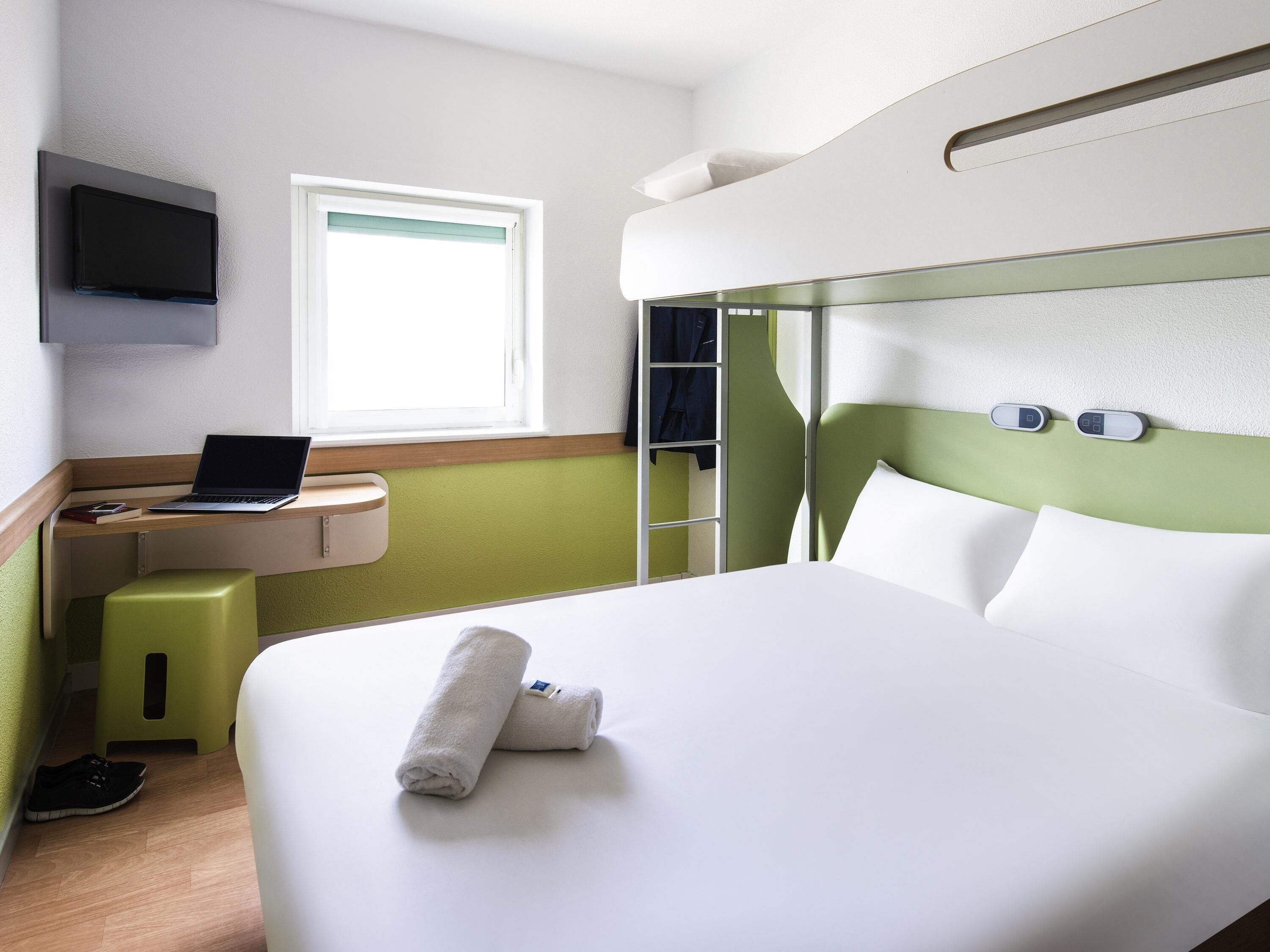 Habitación de hotel con cama, escritorio con computadora portátil, televisor montado en la pared y ventana.