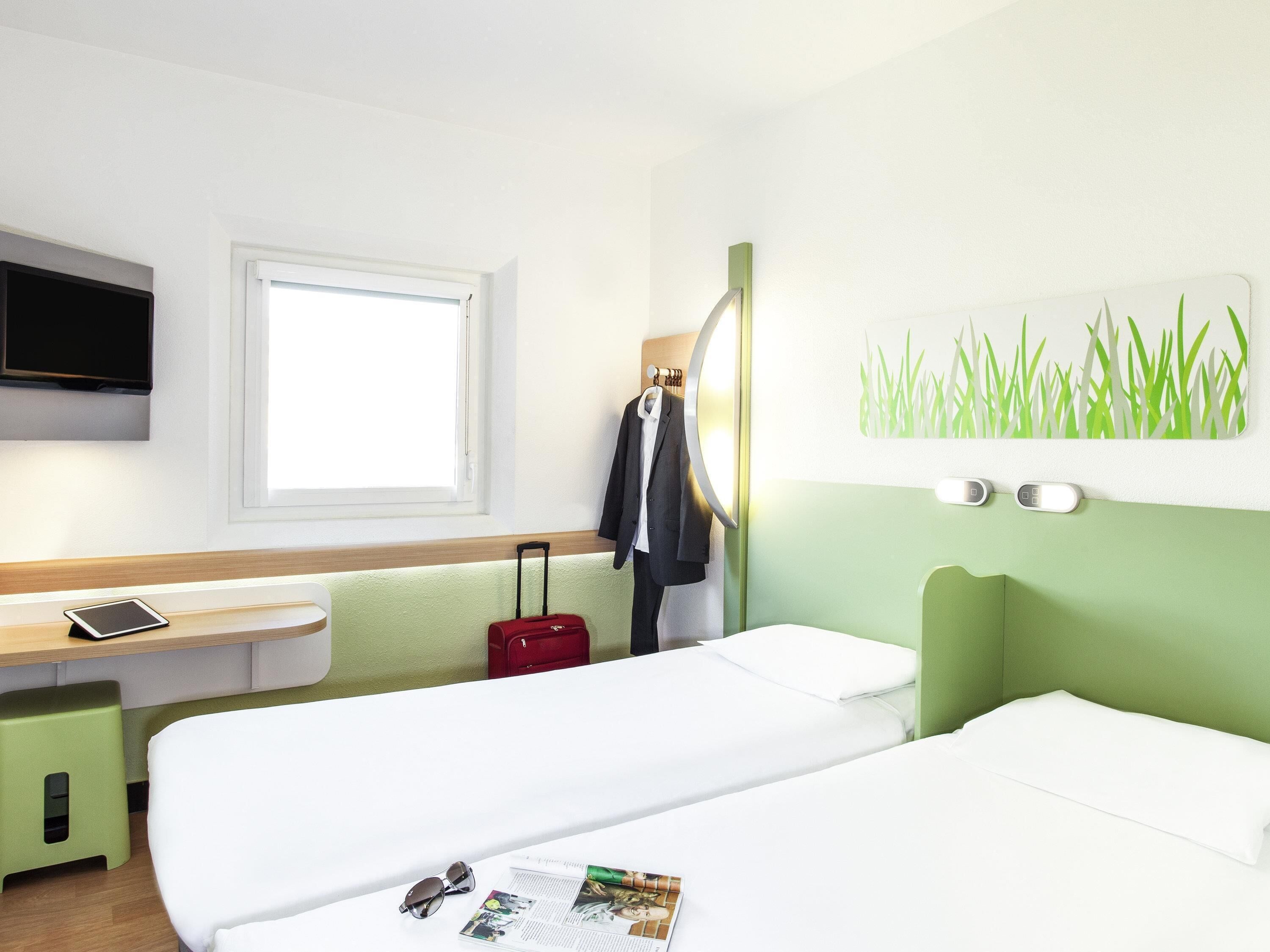 A hotel room with a bed, a desk with a tablet, a red suitcase, a coat on a hook, and a wall-mounted TV.