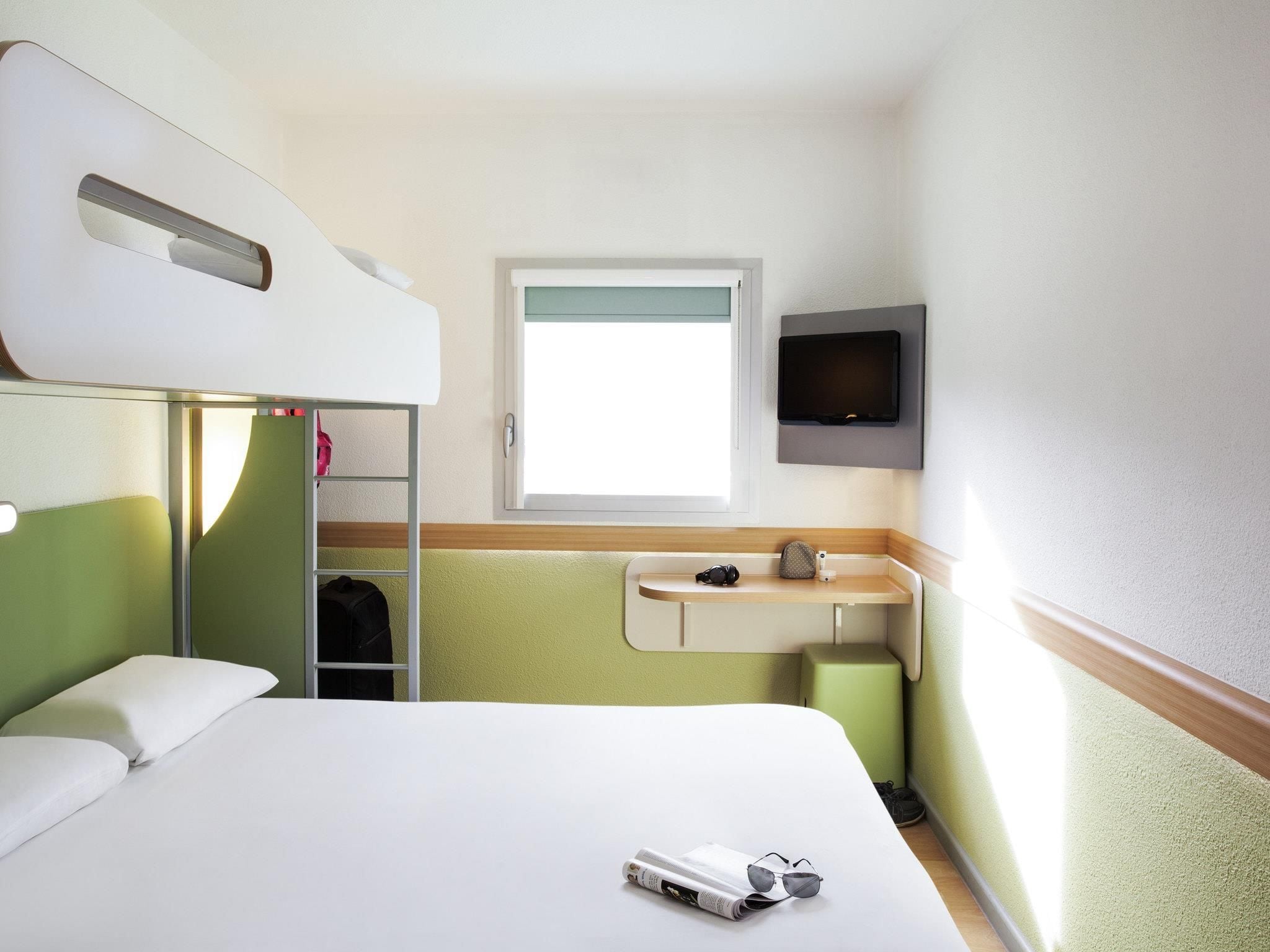 A hotel room with a bunk bed, a small desk with a chair, a television mounted on the wall, and a window with a view.