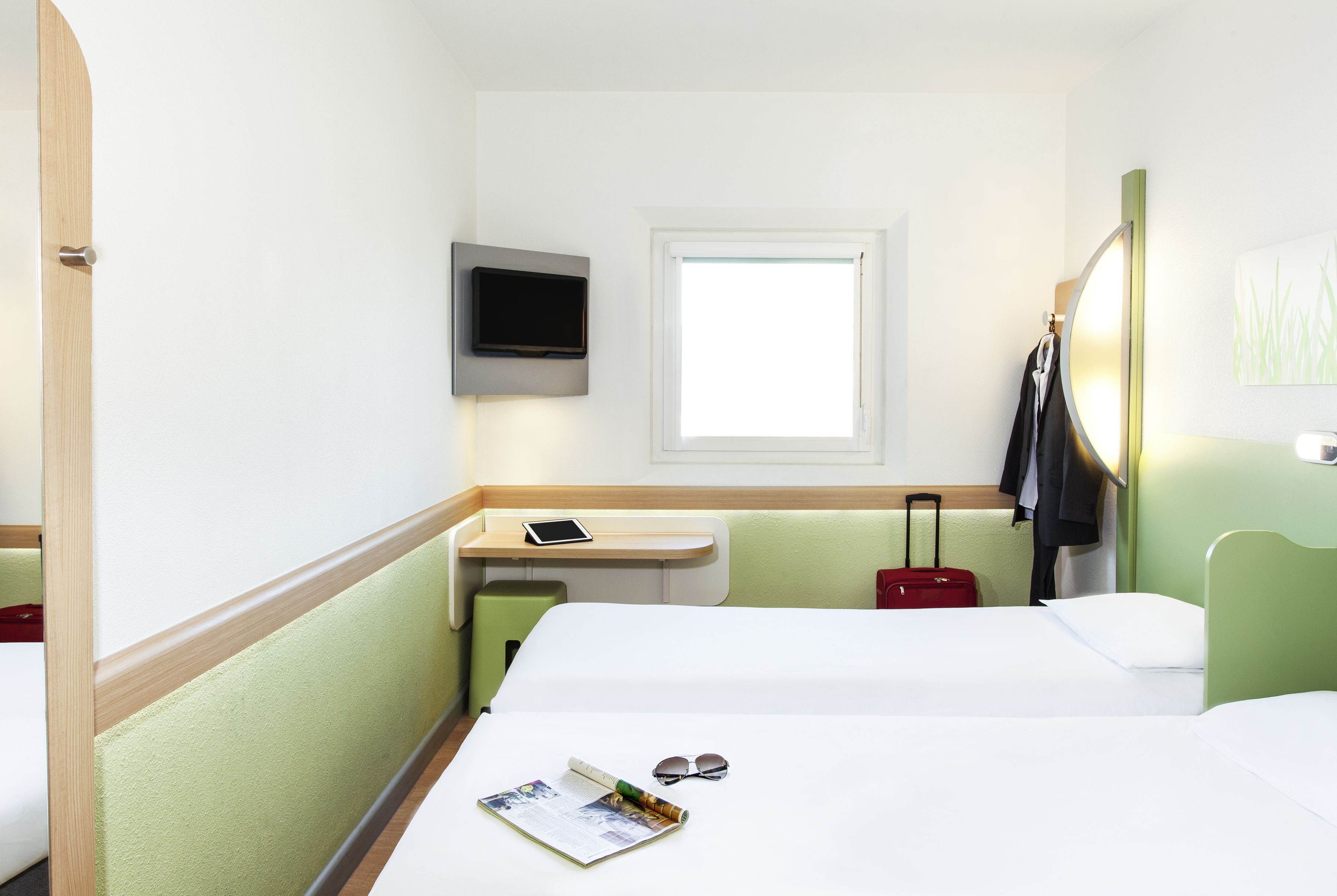 A hotel room with a bed, a desk with a TV, a window, and a suitcase.