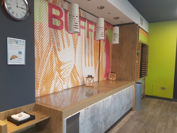 A reception desk with a patterned wall, a clock, and a signboard. - ibis budget London Hounslow (Hounslow)