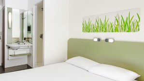 Una habitación de hotel con cama, un baño con lavamanos y espejo, y una pared con un cuadro de un paisaje con hierba verde.