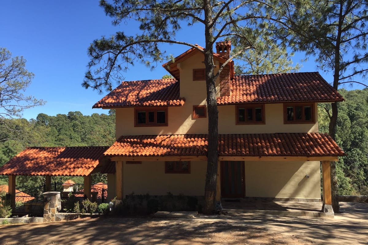 Una casa de dos pisos con techo de tejas rojas, rodeada de árboles y un cielo despejado.