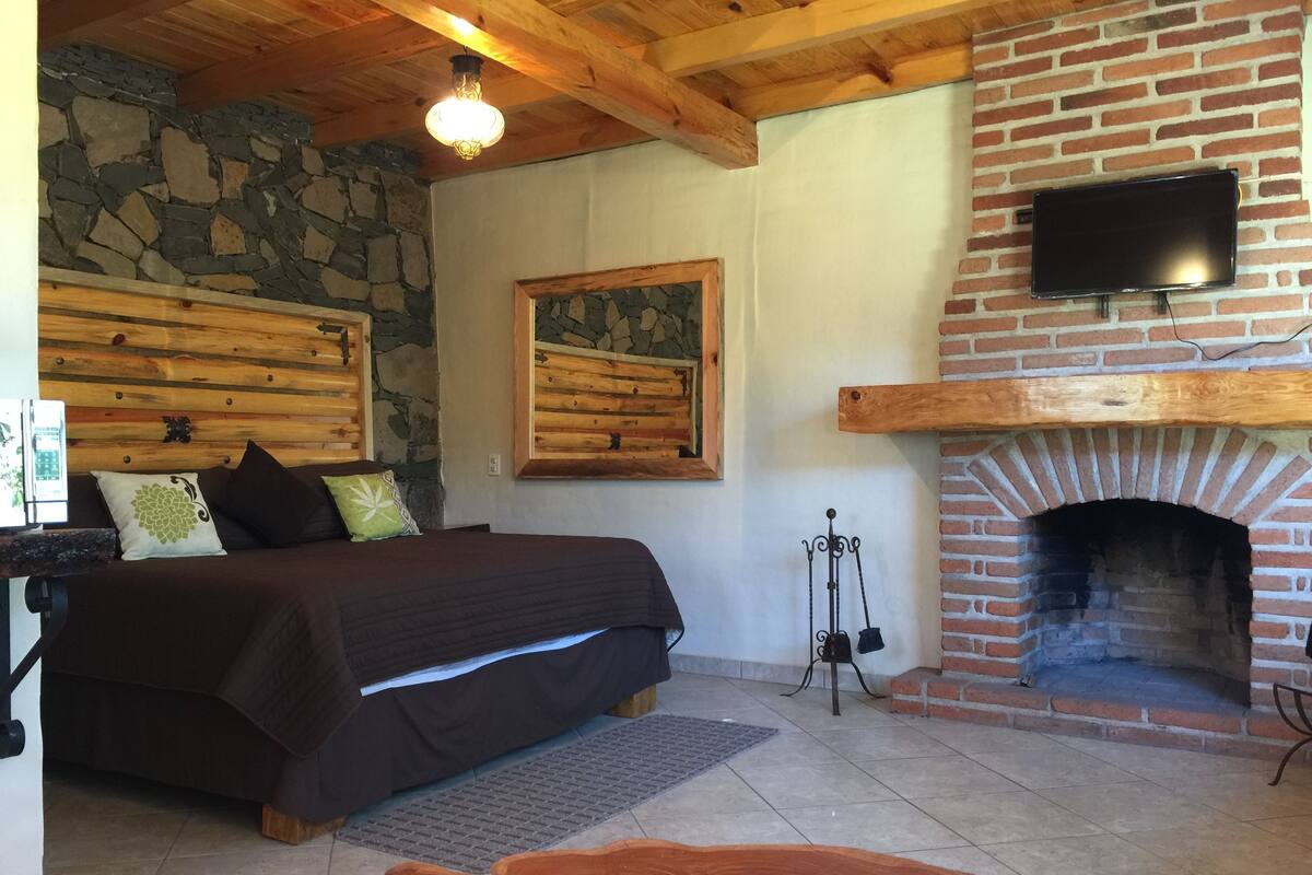 Un dormitorio con pared de piedra, techo de madera, cama con cabecera, chimenea y televisor montado.