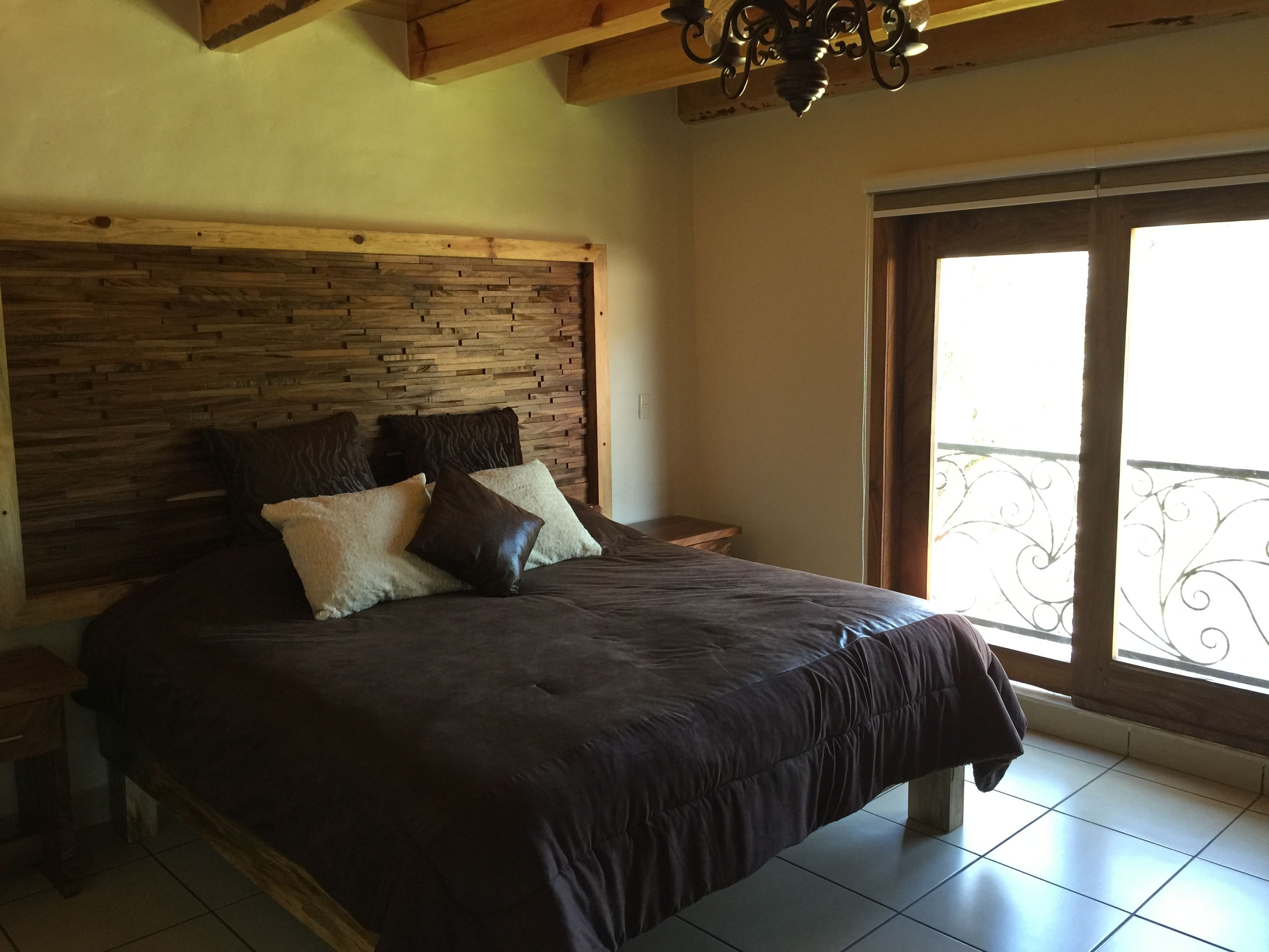 Quarto com cabeceira de madeira, uma cama grande com roupa de cama escura, uma mesinha de cabeceira e uma janela com vista para varanda.