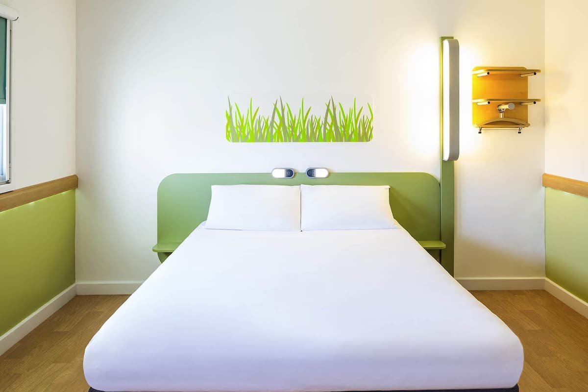 A hotel room with a bed, a wall-mounted light, and a green wall decoration.