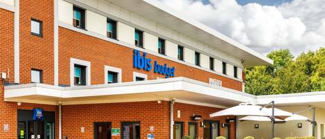 Un hotel de dos pisos con un llamativo letrero de 'ibis budget', una entrada cubierta y un área exterior con jardines.