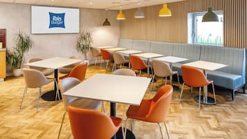 Een moderne vergaderzaal met witte tafels en oranje stoelen, een groot scherm waarop het ibis budget logo staat, en een raam met uitzicht op groen.