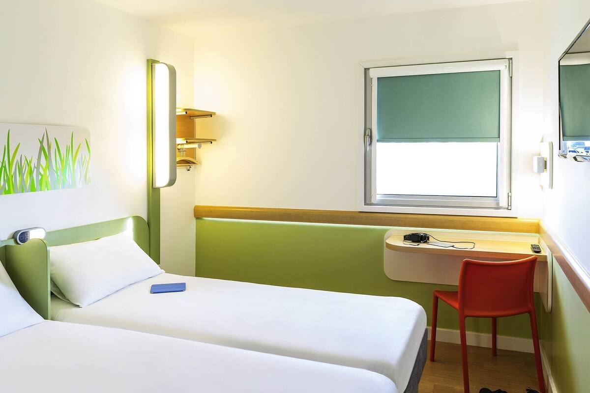 A hotel room with two beds, a desk, a chair, a window, and a wall-mounted mirror.