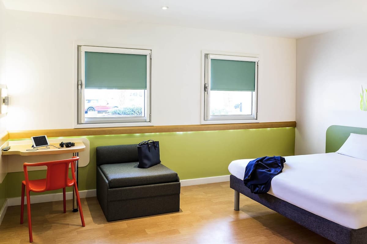 A hotel room with a bed, a desk with a laptop, a chair, a small ottoman, and two windows with blinds.