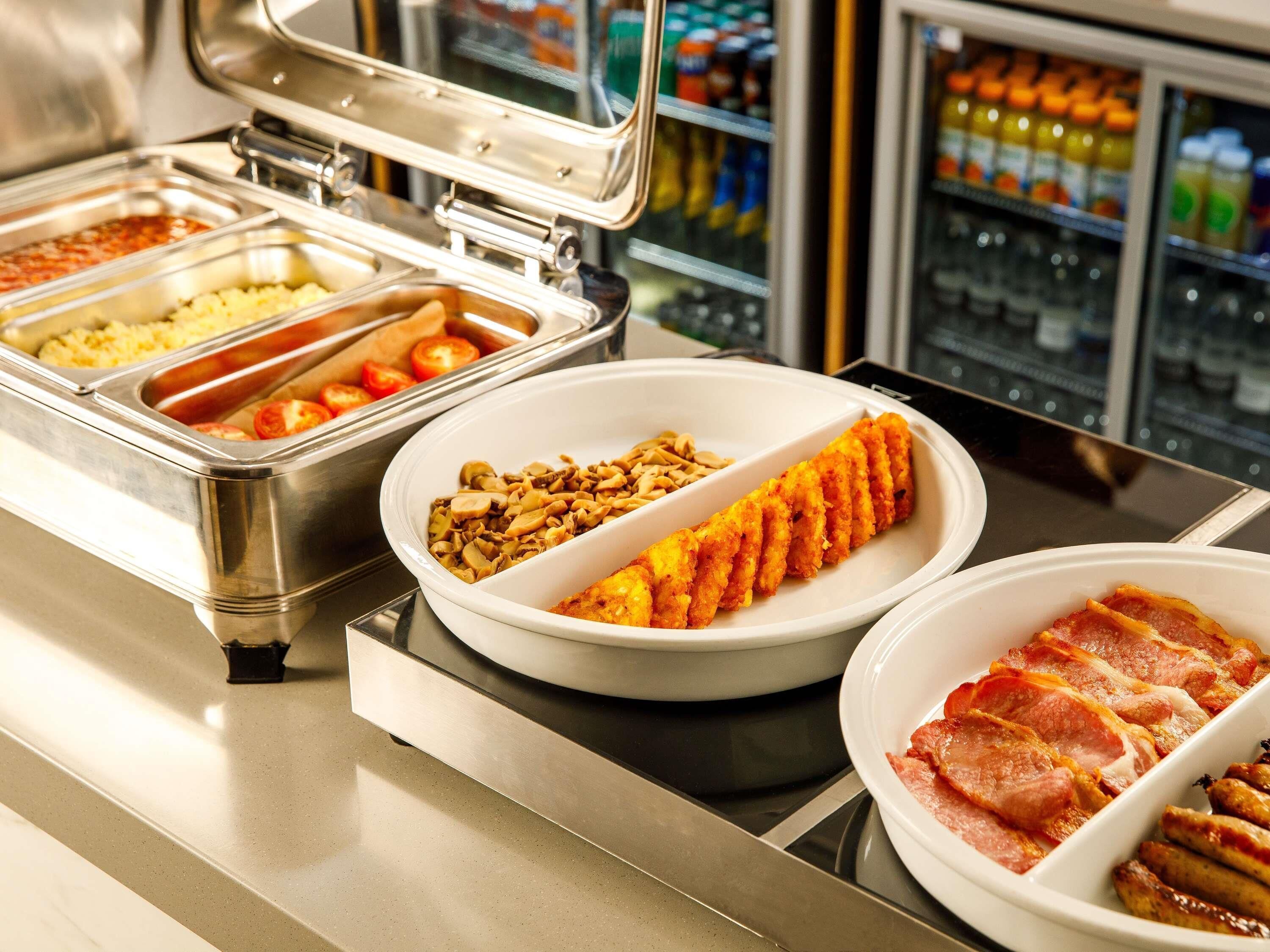 A buffet with various food options including rice, tomatoes, mushrooms, and bacon.