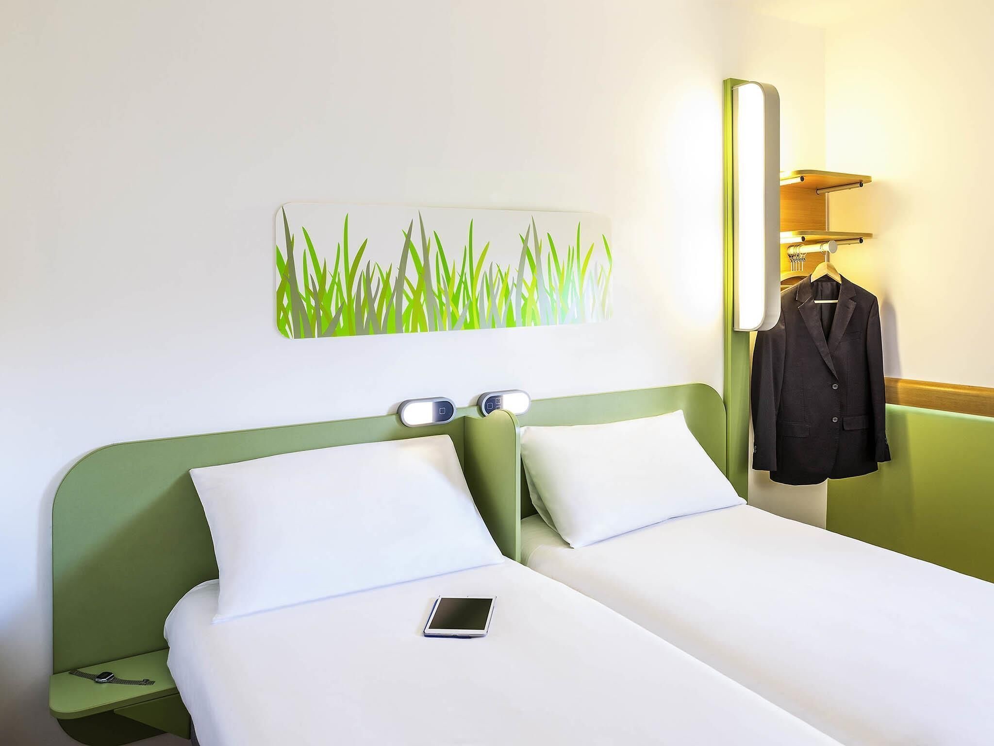 A hotel room with a bed, bedside tables, a wall-mounted lamp, a coat rack, and a painting of grass.