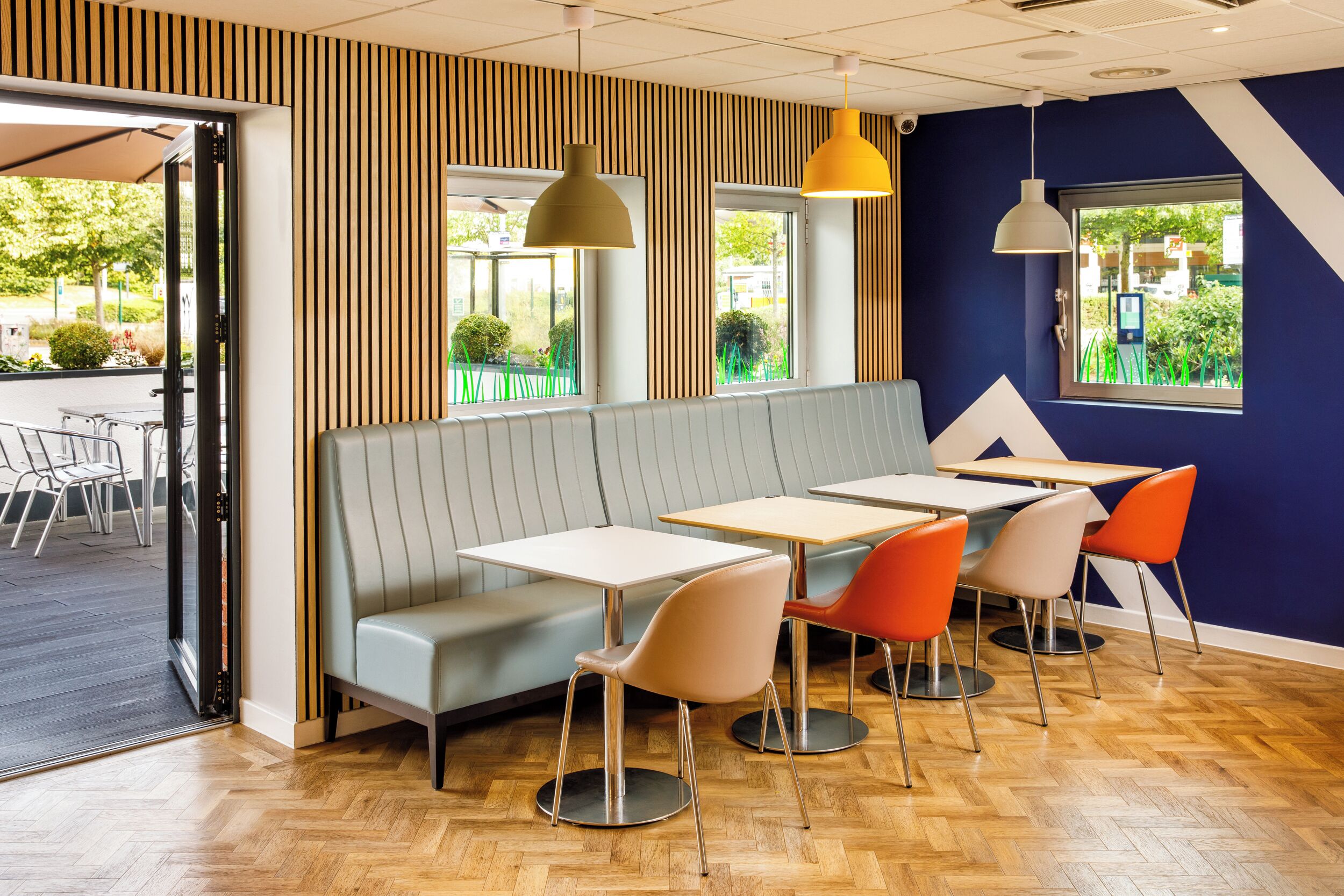 A modern restaurant interior with a wooden herringbone floor, a grey booth seating area, and orange chairs around tables.