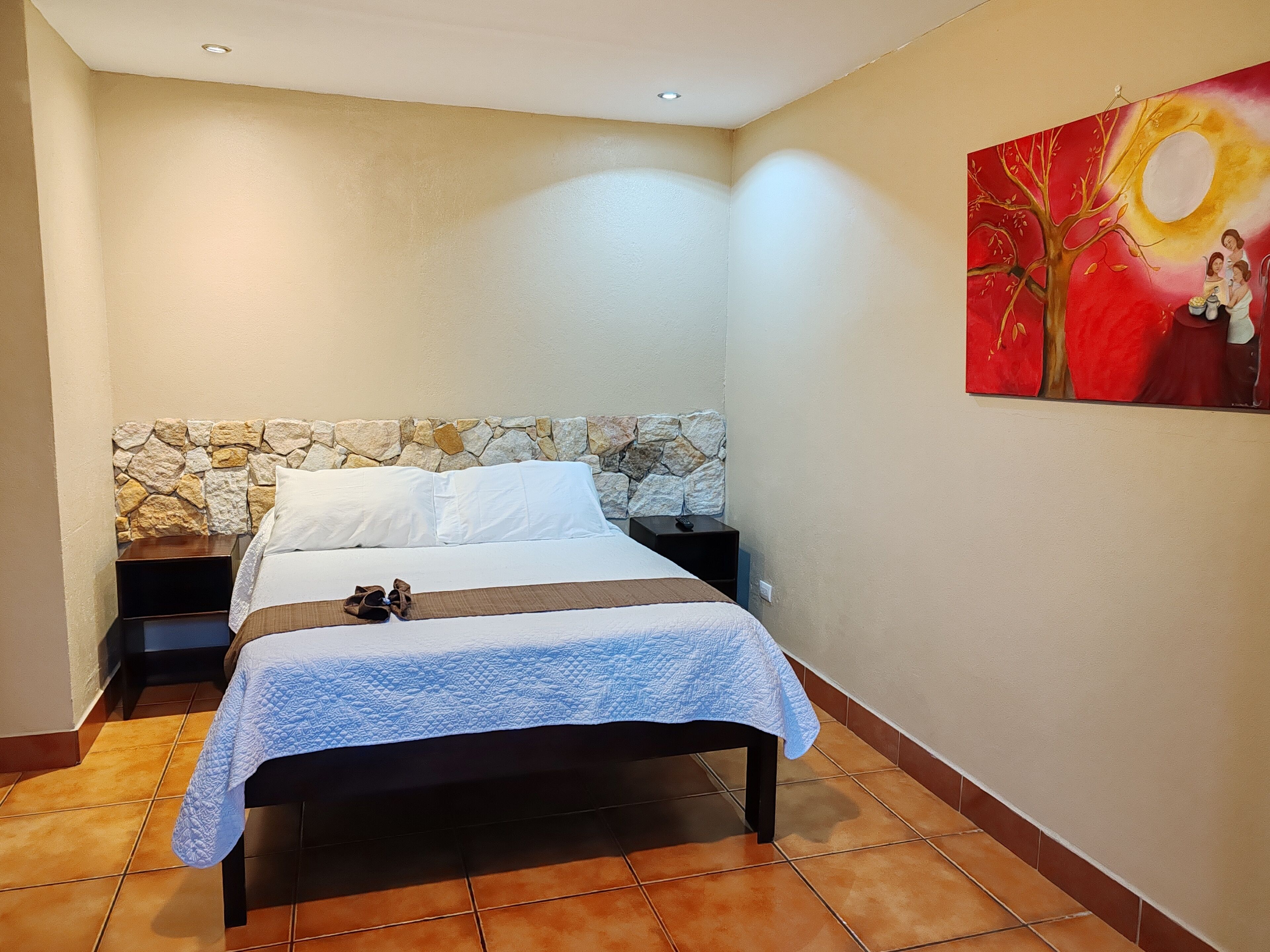 A bedroom with a stone headboard, a bed with a white and blue quilt, a nightstand with a remote control, and a painting on the wall.
