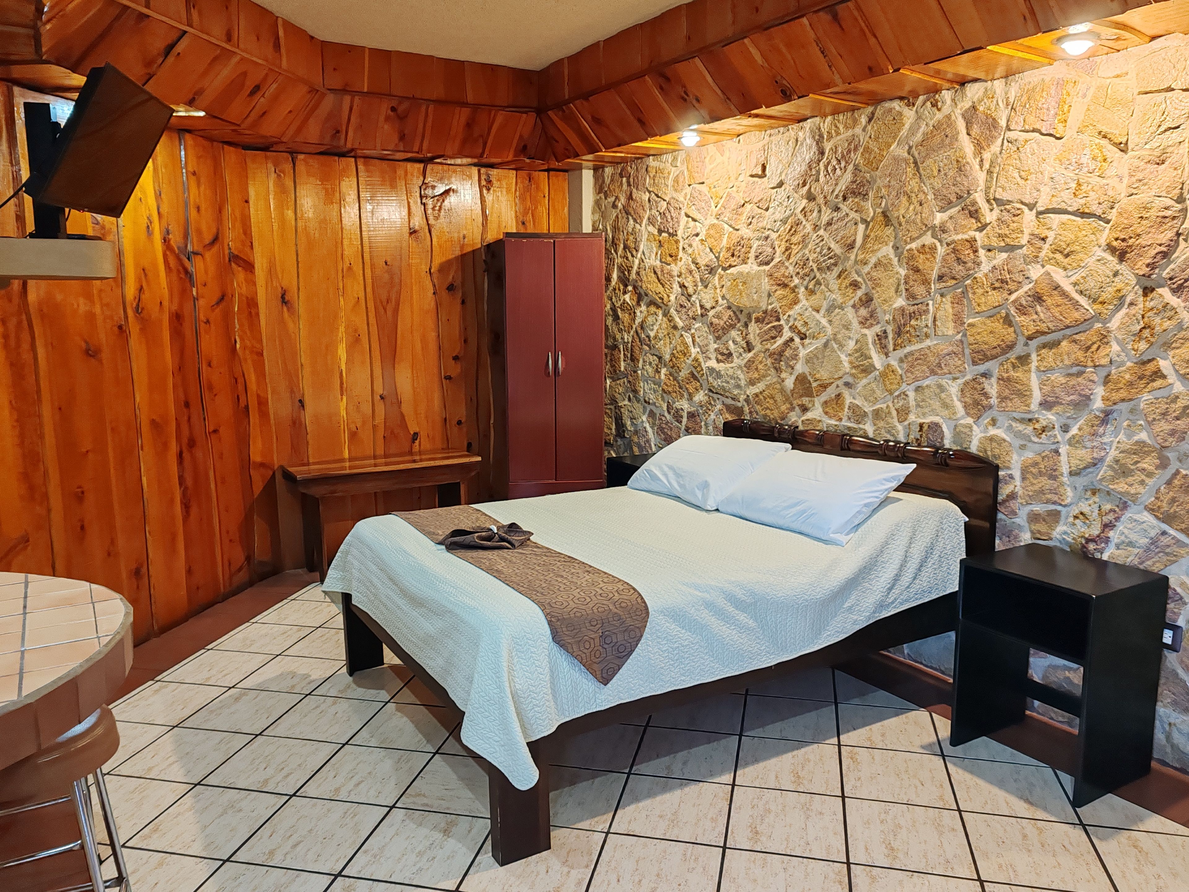 A bedroom with a stone wall, wooden ceiling, a bed with a headboard, a wardrobe, and a bedside table.