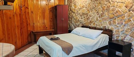 A bedroom with a stone wall, wooden ceiling, a bed with a headboard, a wardrobe, and a bedside table.