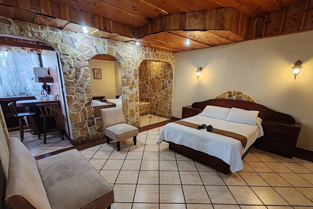 A bedroom with a stone wall, a wooden ceiling, a bed, a chair, and a desk with a chair.