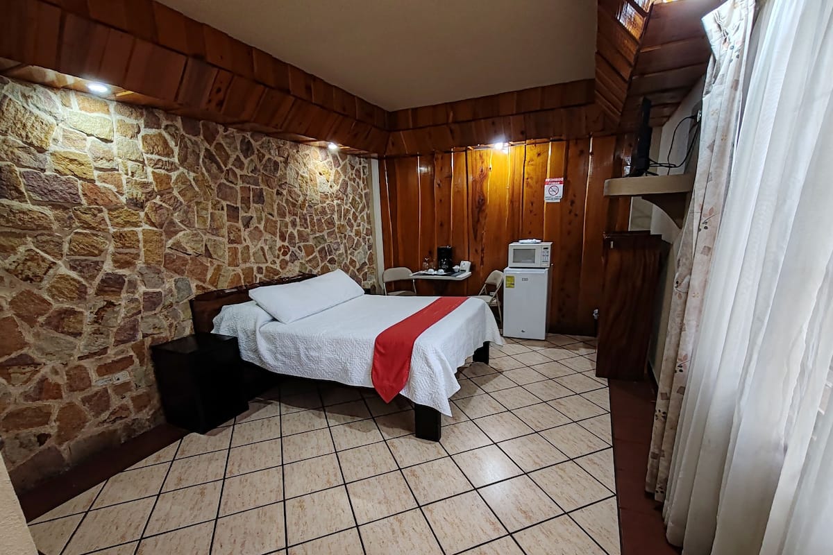 A room with a stone wall, wooden ceiling, a bed with a white bedspread and red blanket, a small desk with a microwave, and a tiled floor.