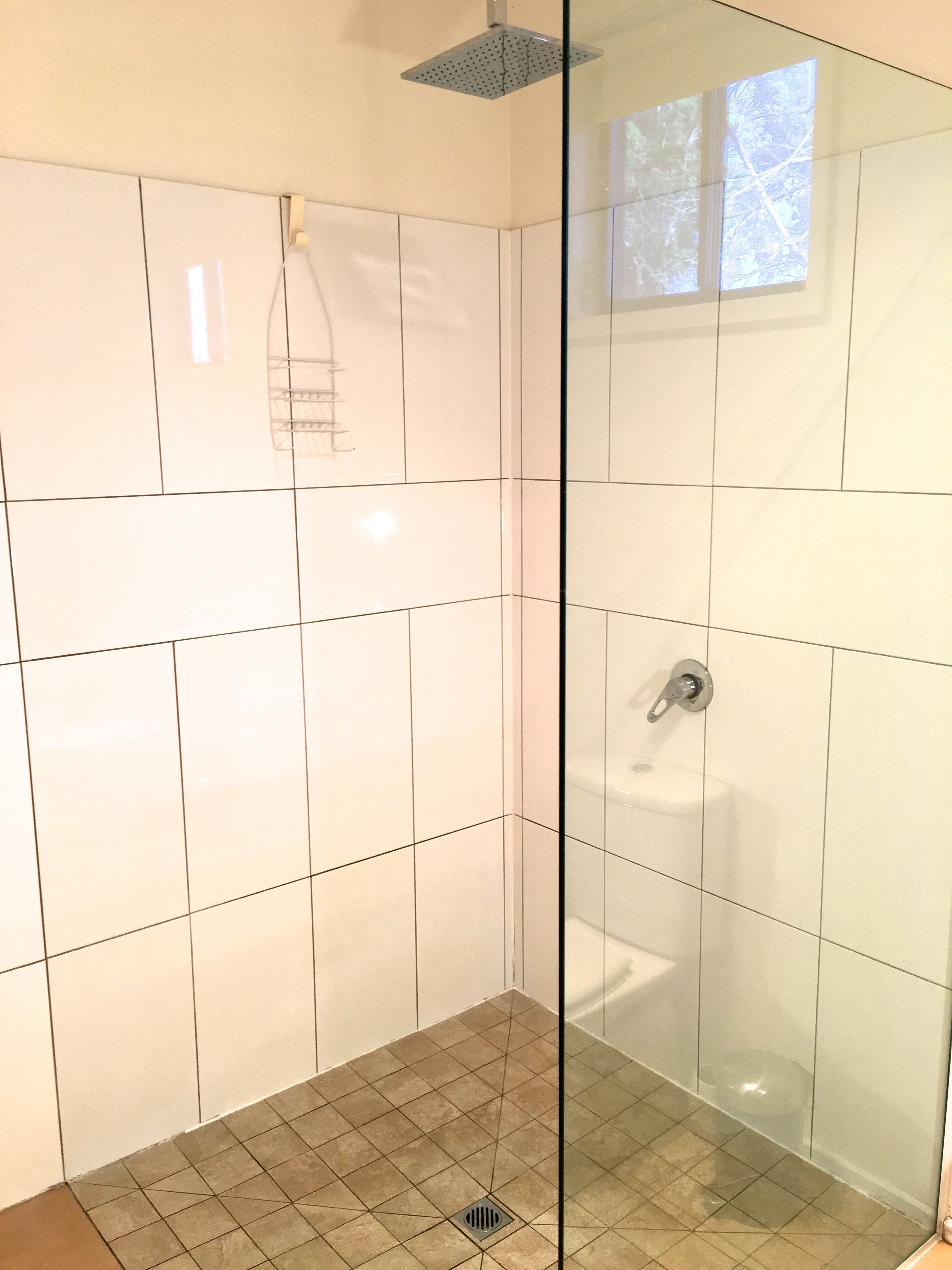 Un bagno con pareti piastrellate in bianco, una zona doccia con porta in vetro e una finestra con vista sugli alberi.