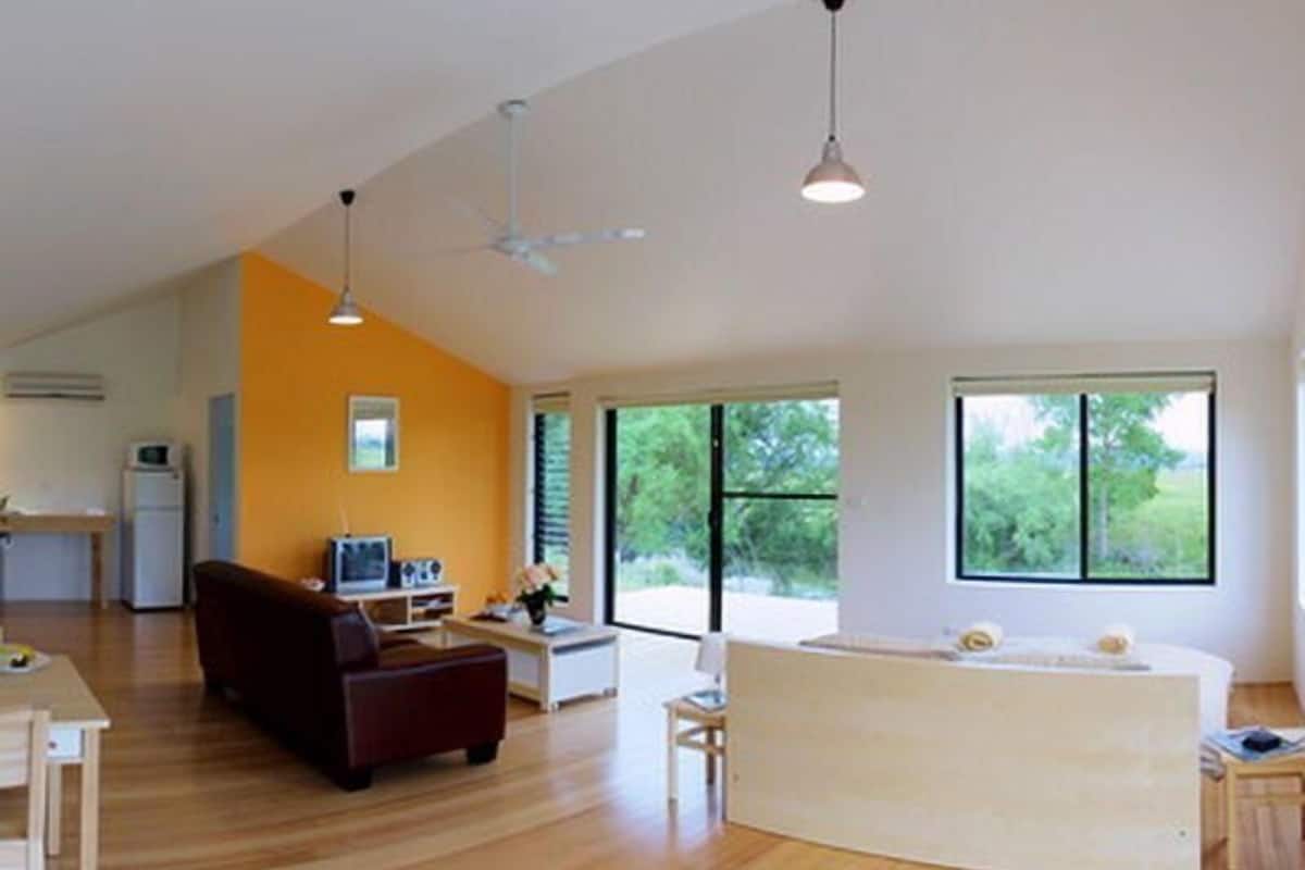 Amplia sala de estar con piso de madera, un sofá, una mesa de centro y un televisor. Amplias ventanas permiten luz natural y ofrecen vistas al exterior.