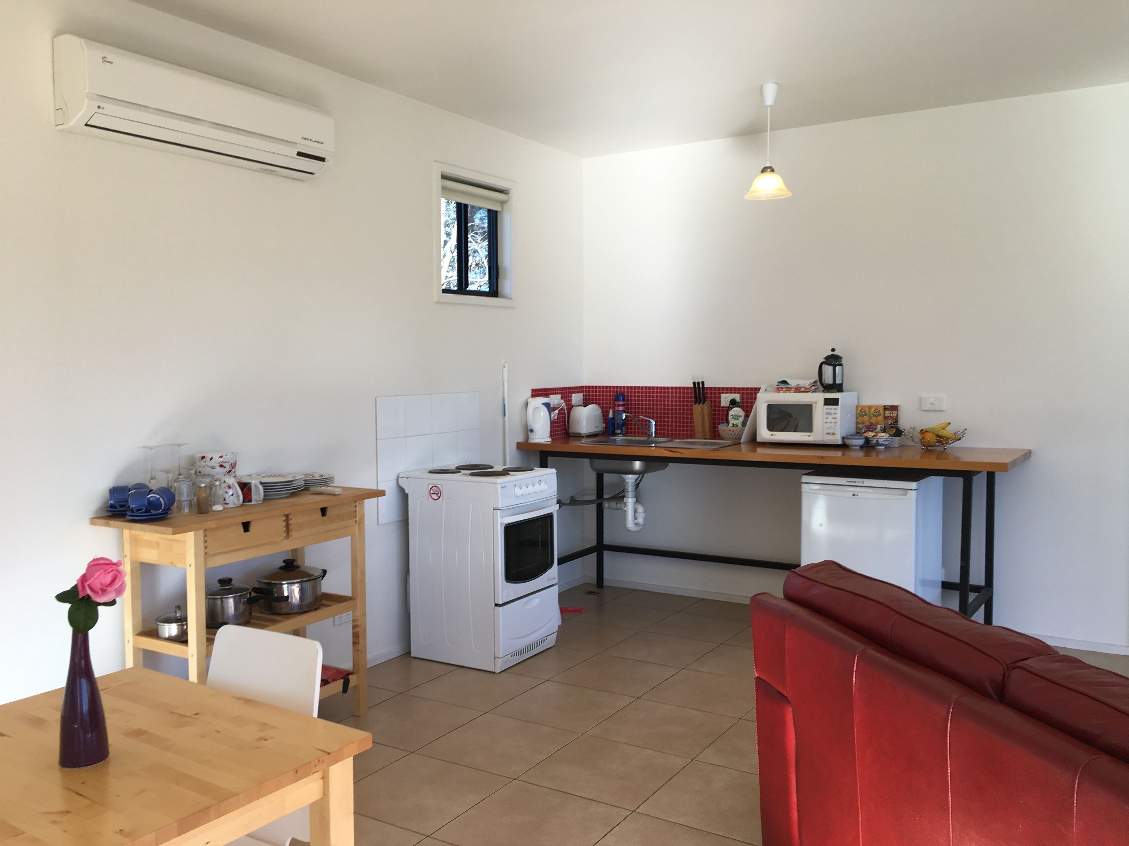 A compact kitchen with a stove, microwave, and sink. There is a wooden table with a vase and a red couch in the foreground.
