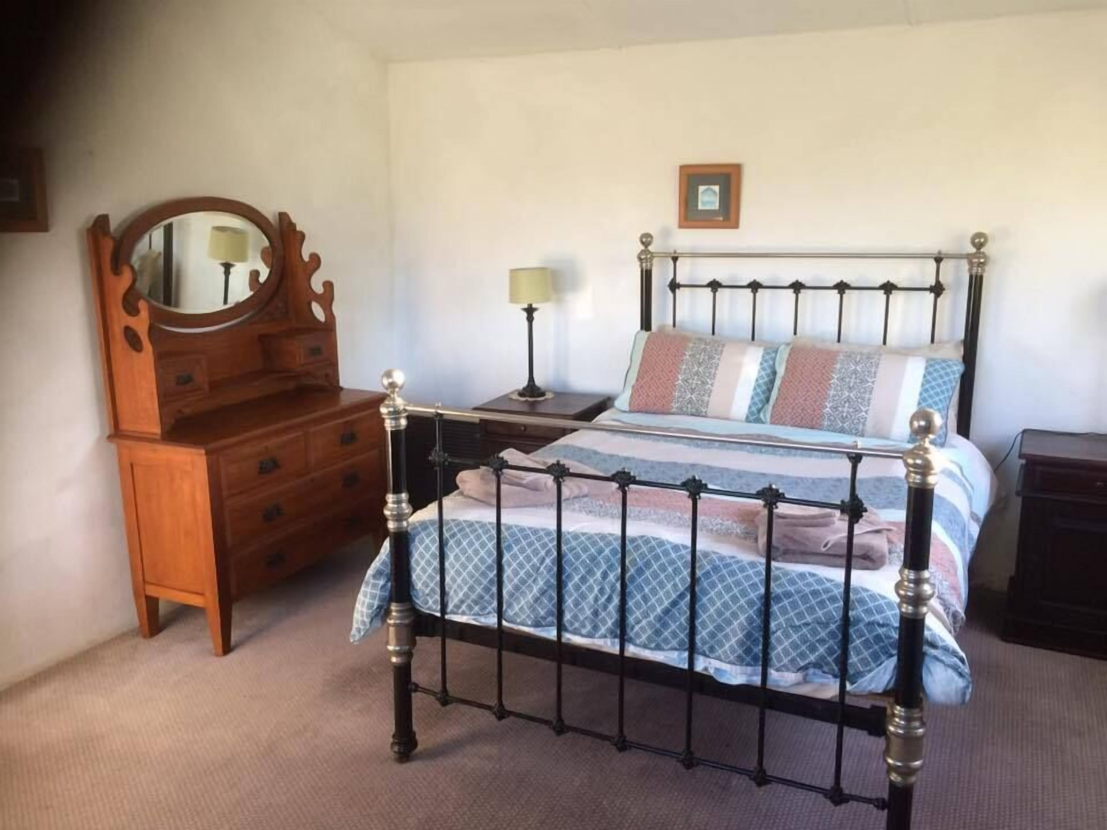 A bedroom with a metal-framed bed, a wooden dresser, a bedside table with a lamp, and a framed picture on the wall.