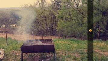 An outdoor grill setup in a wooded area with trees and a dirt path in the background.