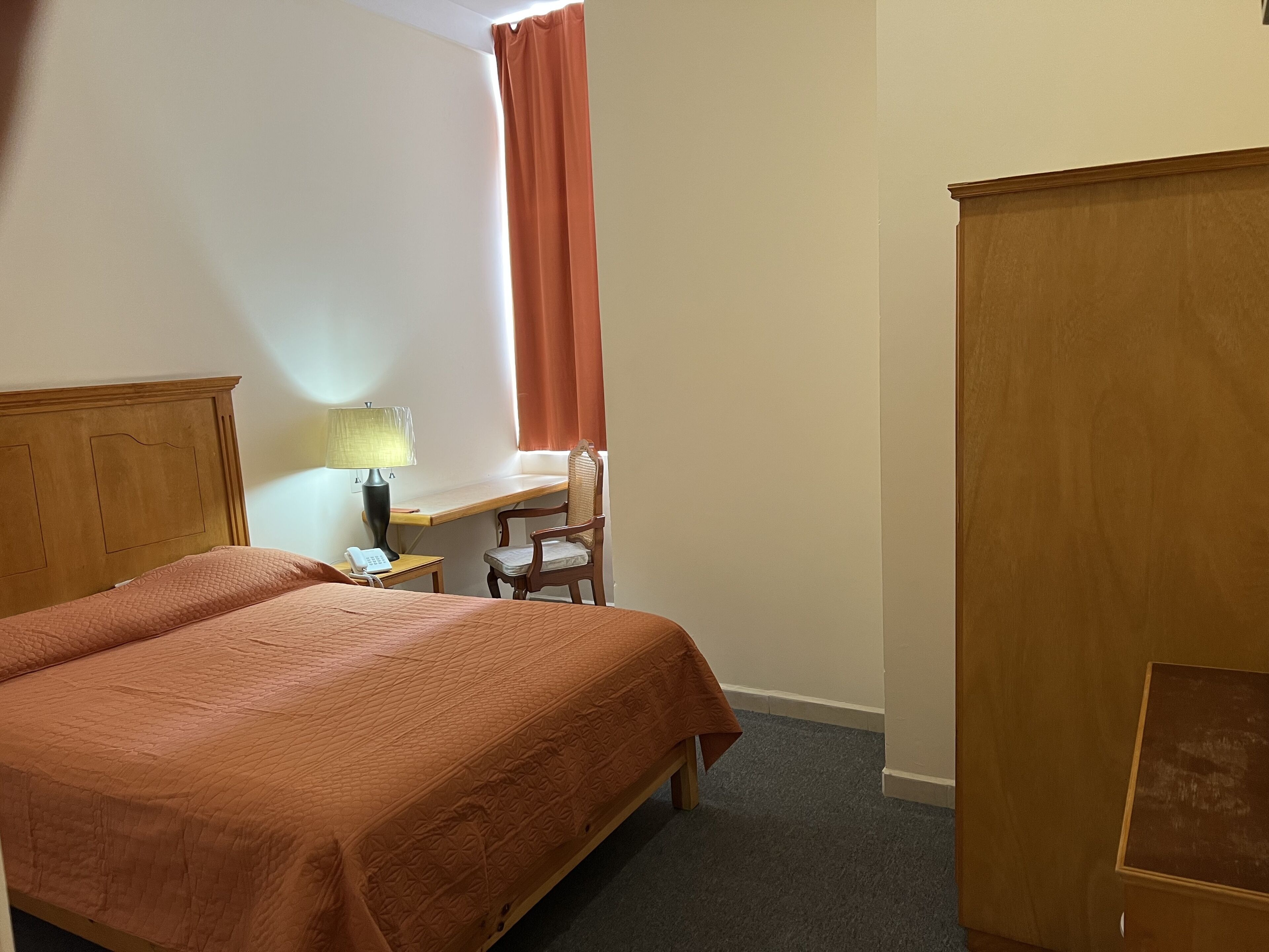 A hotel room with a bed, a desk with a lamp, a chair, and a wardrobe.