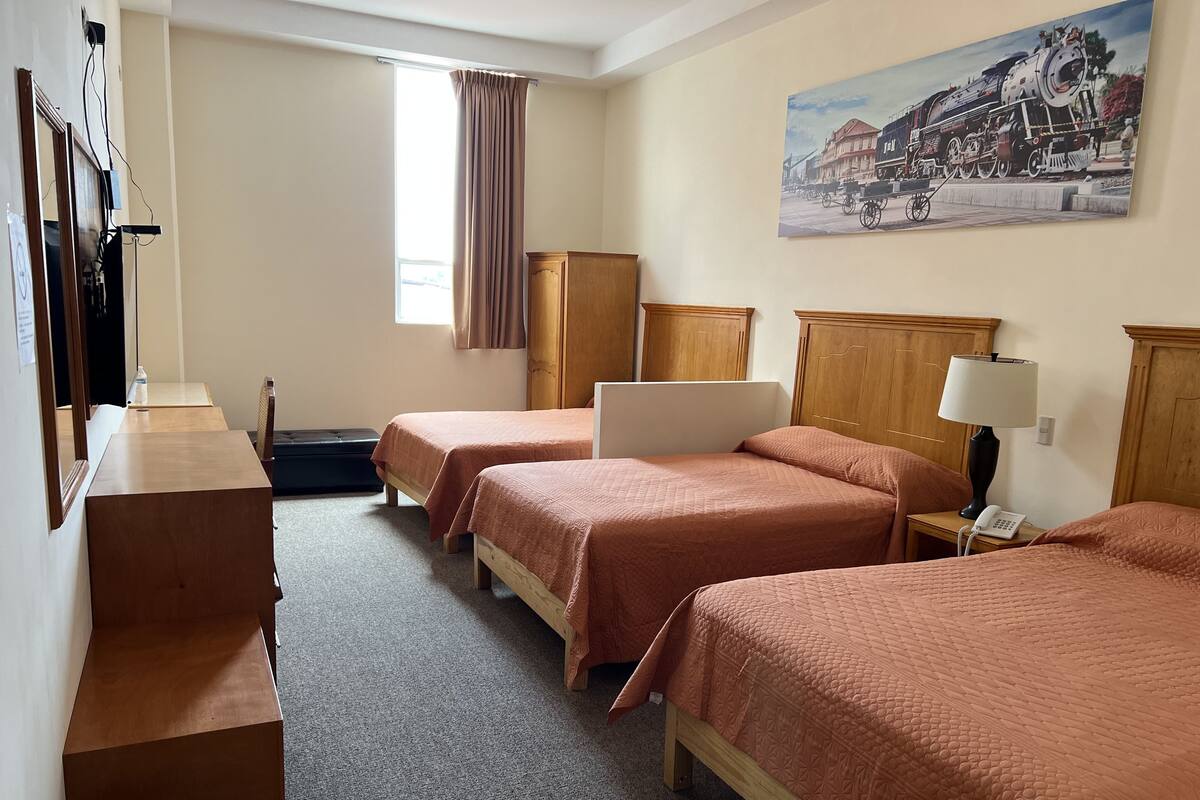 Une chambre d’hôtel avec trois lits, un bureau, une chaise, une poubelle, une fenêtre avec des rideaux et un tableau au mur.