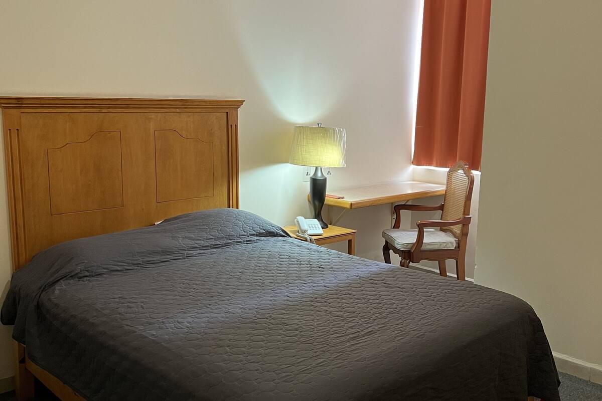 Une chambre à coucher avec un lit en bois, un bureau avec une lampe, une chaise et une fenêtre avec des rideaux.