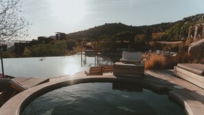 Un área de piscina con sillones de descanso y un jacuzzi, rodeado de paisaje natural y una colina.