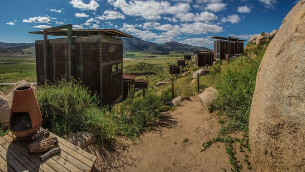 Un hotel de estilo rústico tipo cabaña, con varias cabañas de madera, un sendero de tierra que atraviesa un paisaje montañoso y un cielo azul despejado.