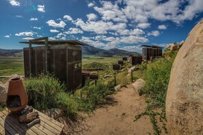 Un hotel de estilo rústico tipo cabaña, con varias cabañas de madera, un sendero de tierra que atraviesa un paisaje montañoso y un cielo azul despejado.