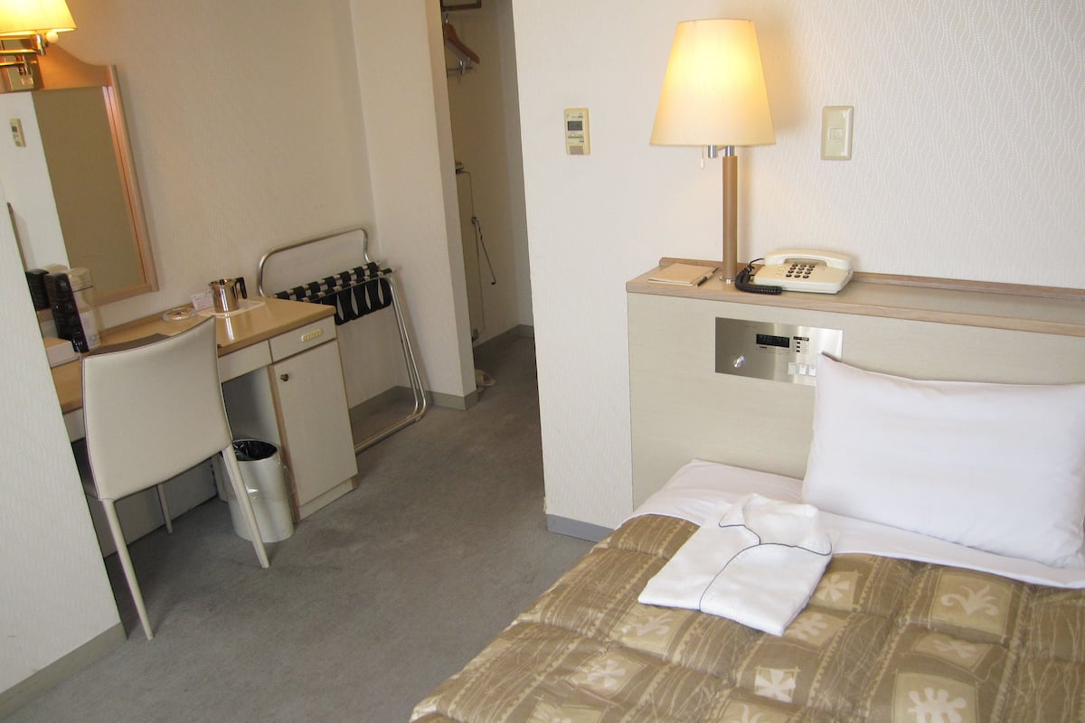 A hotel room with a bed, a desk, a chair, a telephone, and a lamp.