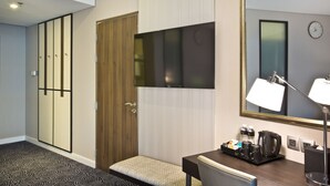 Habitación de hotel con armario de madera, televisor de pantalla plana, escritorio con silla, banca, espejo y bandeja con artículos de aseo.