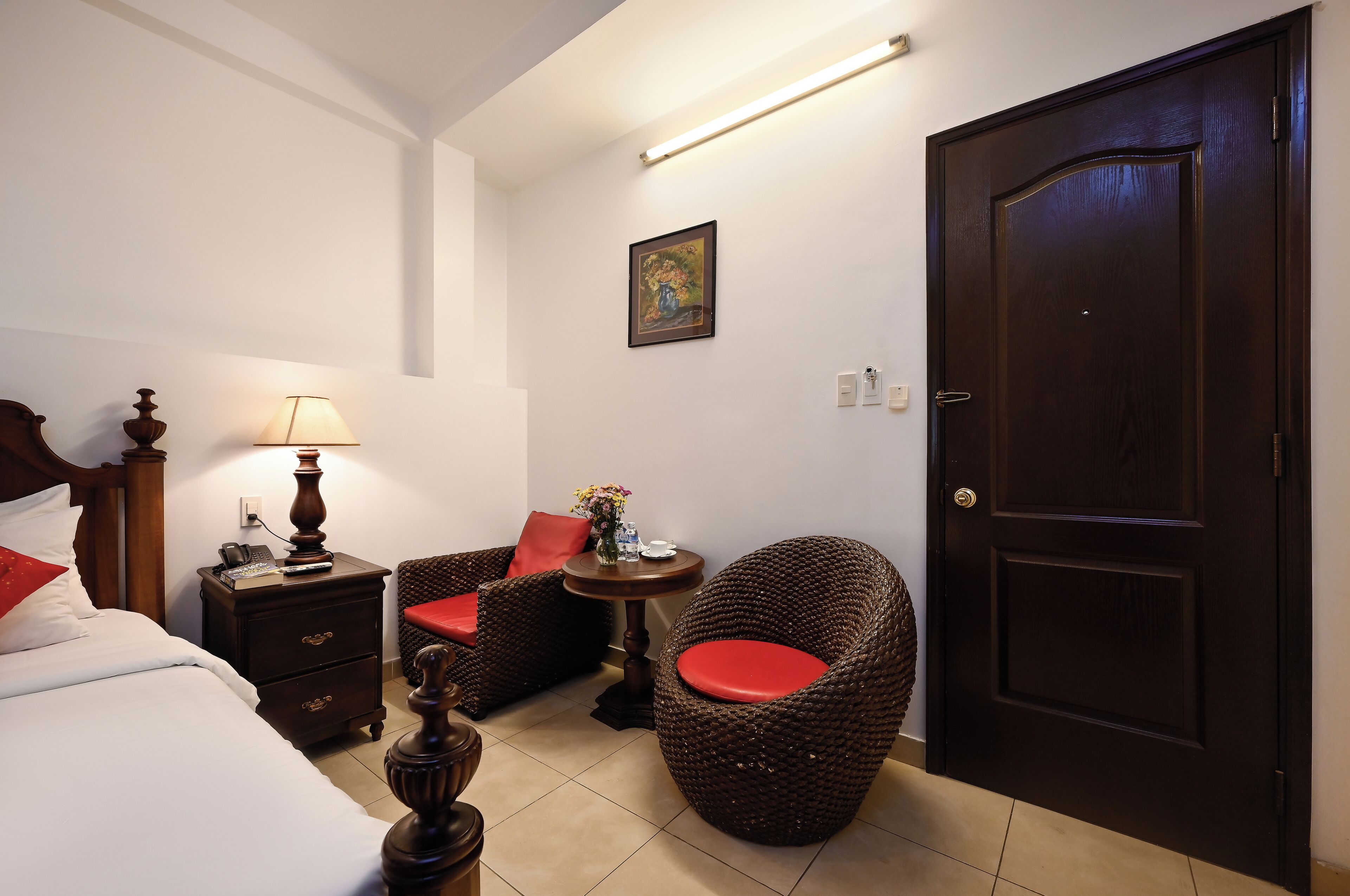 A hotel room with a bed, a nightstand, a lamp, a wicker chair with a red cushion, a small table with a vase of flowers, and a dark wooden door.