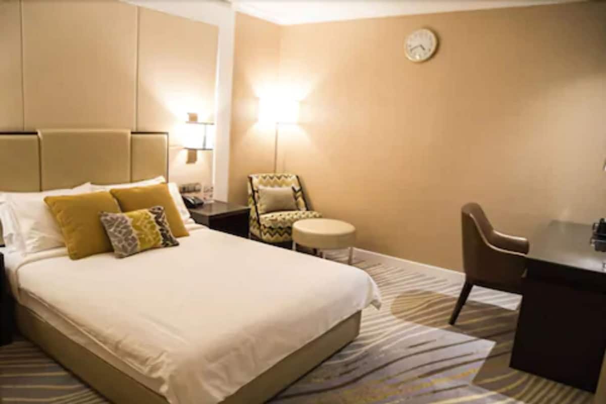 A hotel room with a large bed, a desk, a chair, a small table, and a lamp.