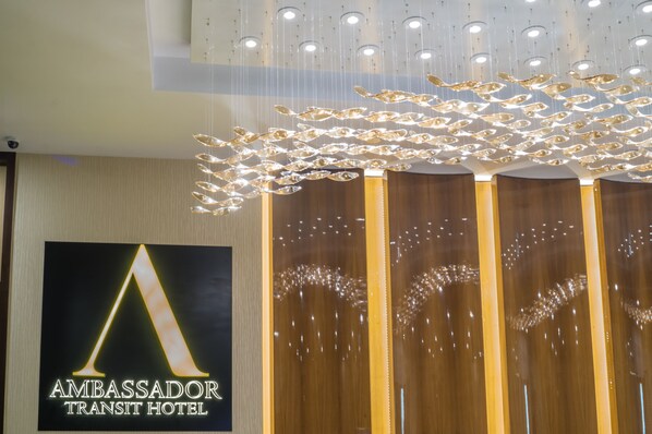 A hotel reception area with a prominent 'Ambassador Transit Hotel' sign, modern lighting fixtures, and a wooden reception desk. - Ambassador Transit Hotel Terminal 2 (Singapore)