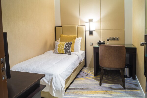 A hotel room with a bed, a desk, a chair, a lamp, and a trash bin.