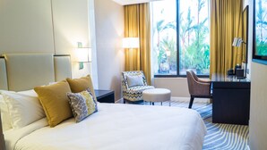Habitación de hotel con una cama grande, un escritorio, una silla y vistas a palmeras a través de la ventana.