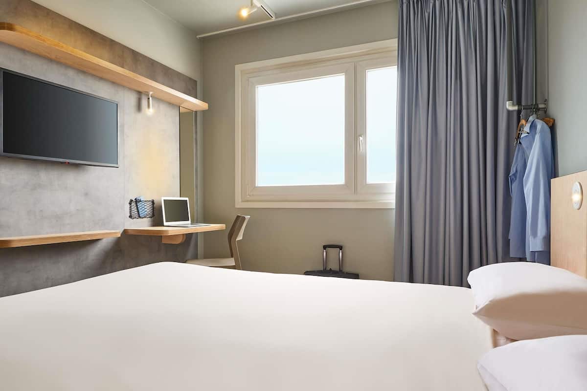 A hotel room with a bed, a desk with a laptop, a TV, a window with curtains, and a suitcase.