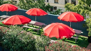 Terrasse extérieure avec des parasols rouges, entourée de verdure et avec un bâtiment en arrière-plan.