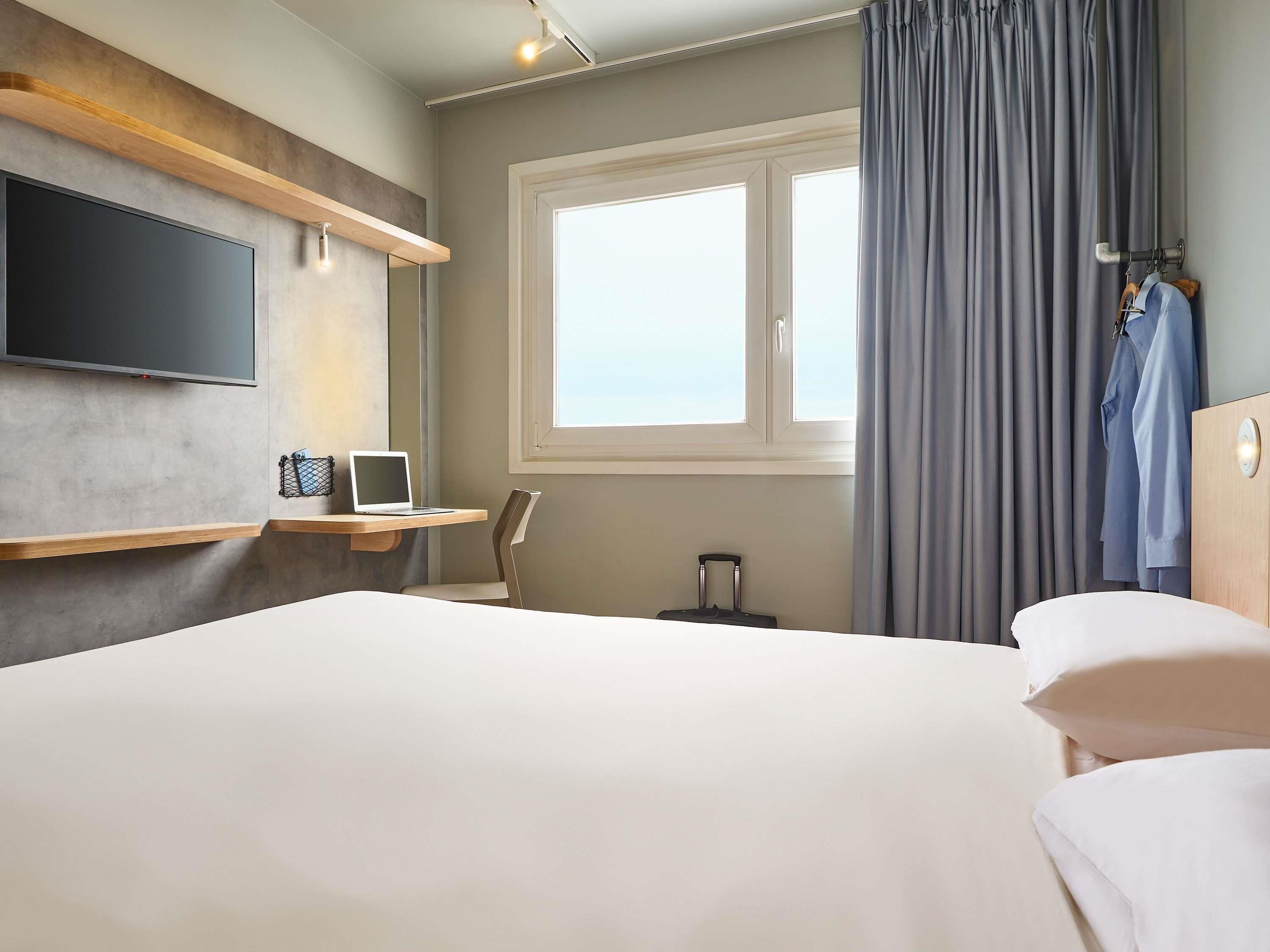 Une chambre d’hôtel avec un lit, un bureau avec un ordinateur portable, une télévision, une fenêtre avec des rideaux et une valise.