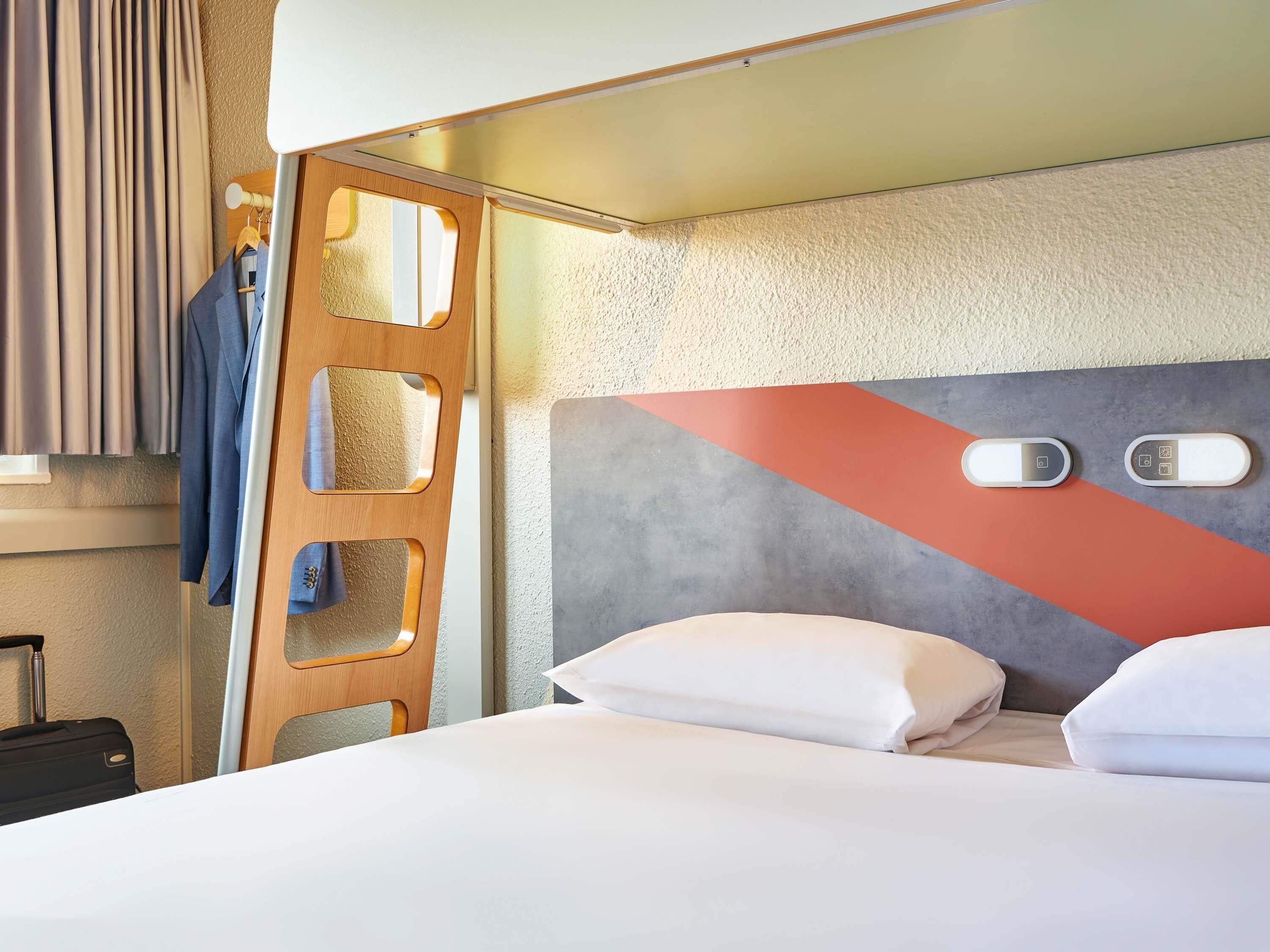 A hotel room with a bed, pillows, a wooden wardrobe, and a coat rack with clothes.