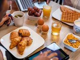 A breakfast setting with croissants, eggs, a waffle, and a laptop on a wooden table.