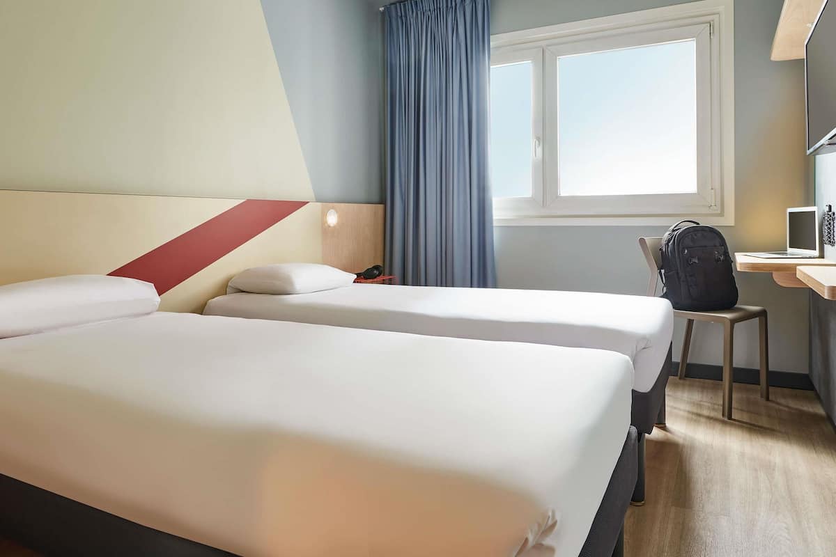A hotel room with two beds, a desk with a laptop, and a window with curtains.