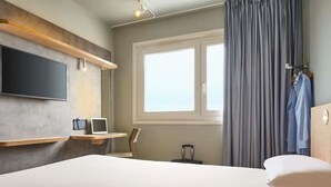 Une chambre d’hôtel avec un lit, un téléviseur fixé au mur, une fenêtre avec des rideaux, un bureau avec une chaise et une valise.