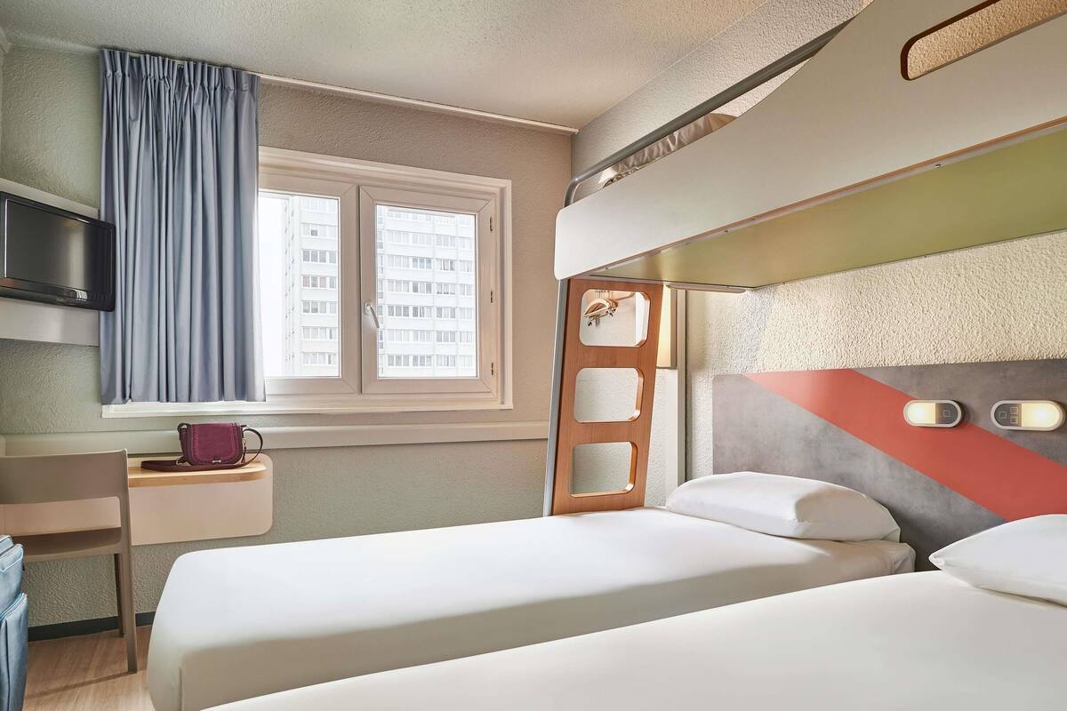 A hotel room with two beds, a desk, a chair, a TV, and a window with curtains.